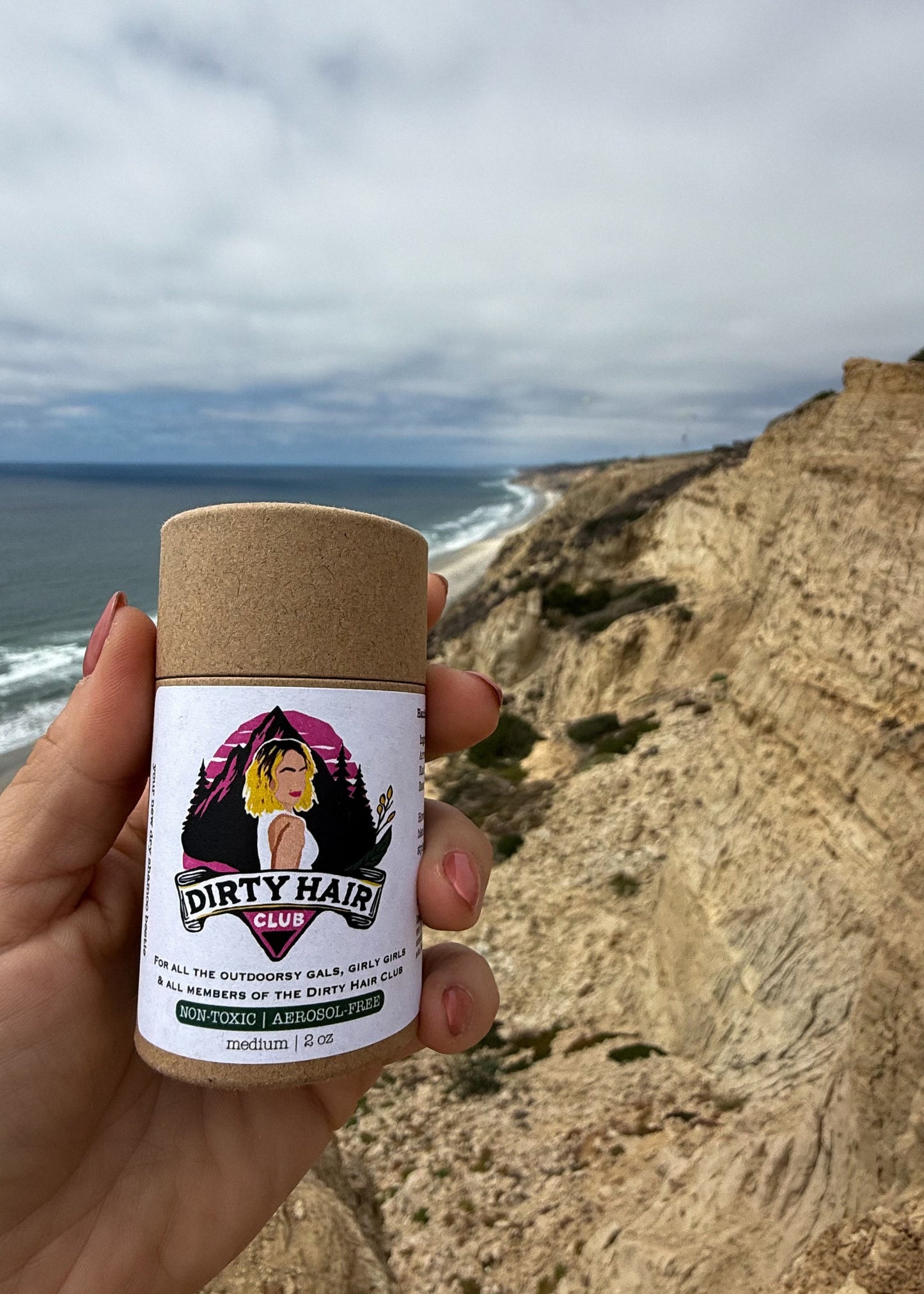 Hand holding a 'Dirty Hair club dry shampoo product container with a coastal background