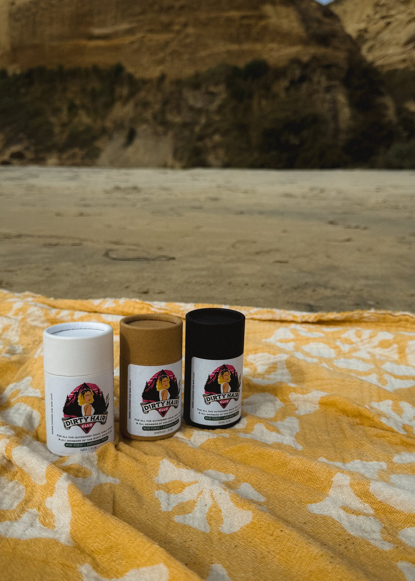 Three cylindrical containers dry shampoo product on a yellow blanket against a desert background