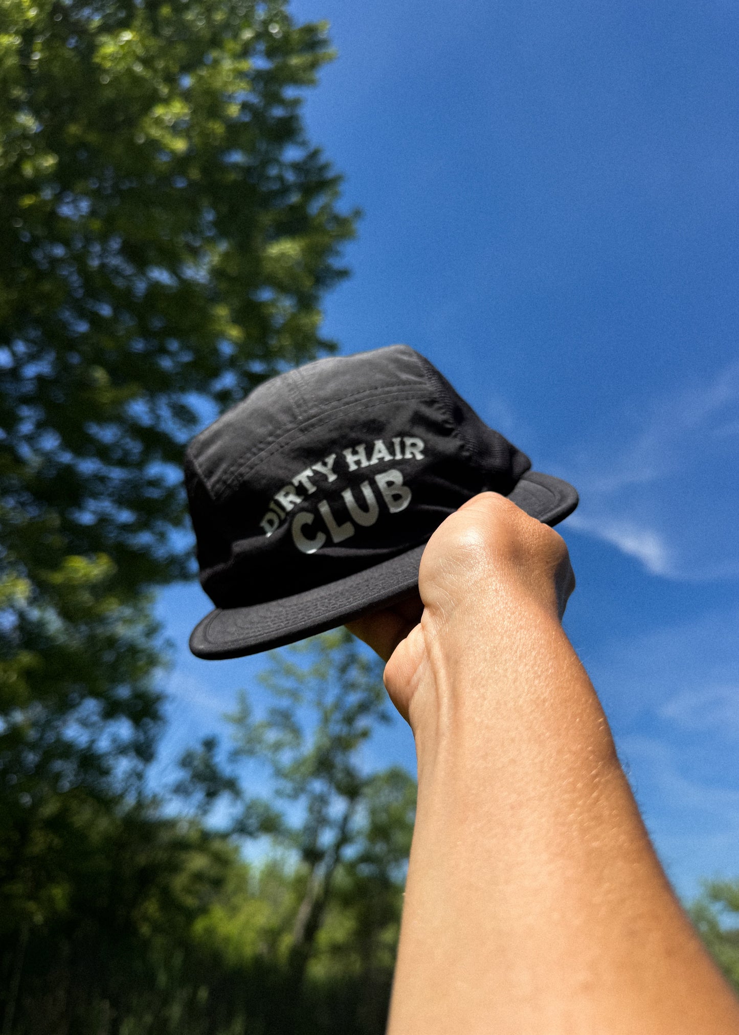 Black cap with 'Dirty Hair Club' text held against a blue sky with trees