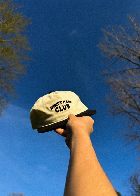 Hand holding a beige cap with 'Dirty Hair Club' text against a blue sky.