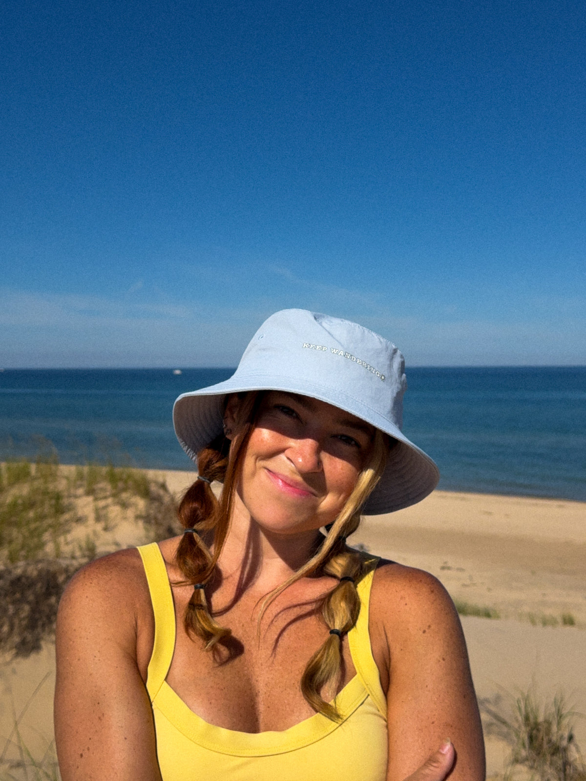 A woman wearing a light blue wide-brim sun hat and a yellow tank top stands on a sandy beach with the ocean and blue sky in the background.