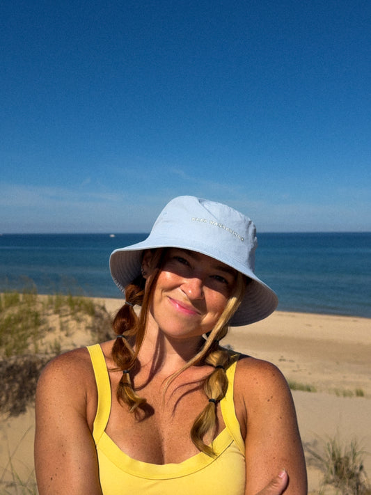 A woman wearing a light blue wide-brim sun hat and a yellow tank top stands on a sandy beach with the ocean and blue sky in the background.