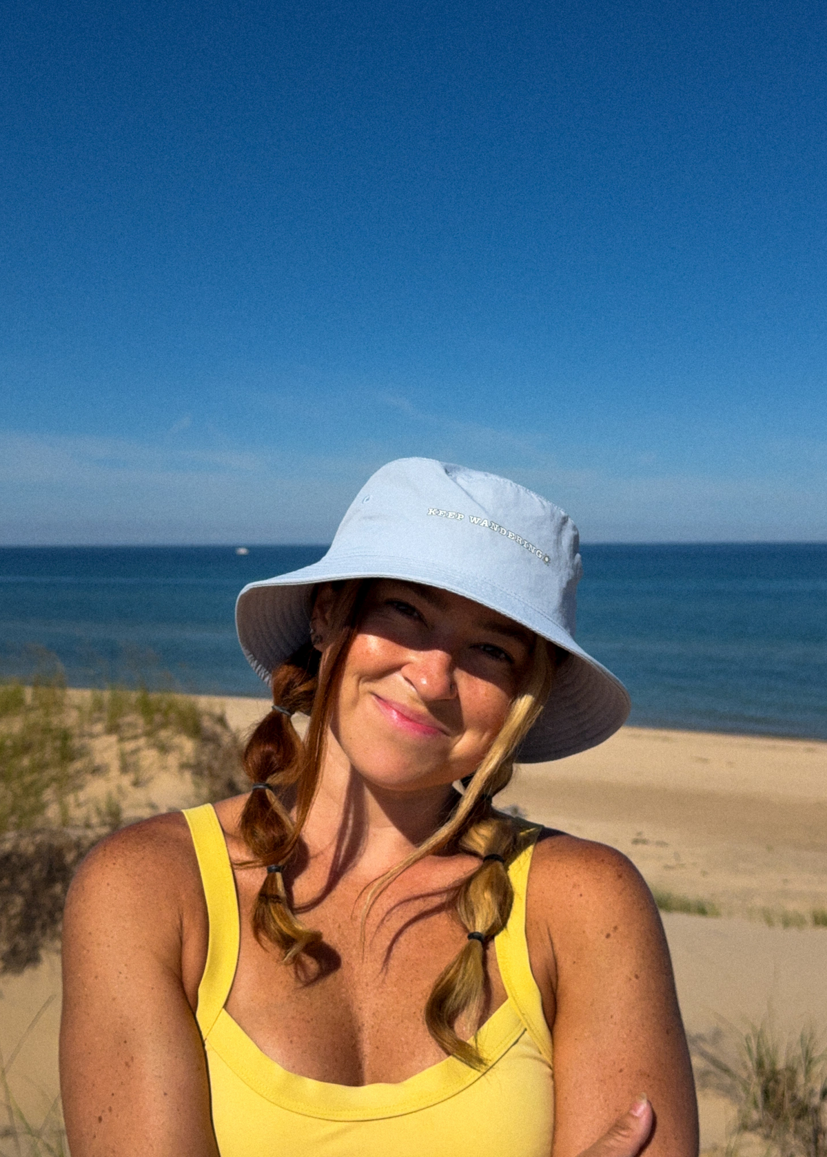 A woman on a beach wearing a light blue wide-brim sun hat and a yellow tank top, with the ocean and sand in the background.