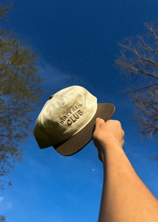 Hand holding a beige cap with 'Dirty Hair Club' text against a blue sky