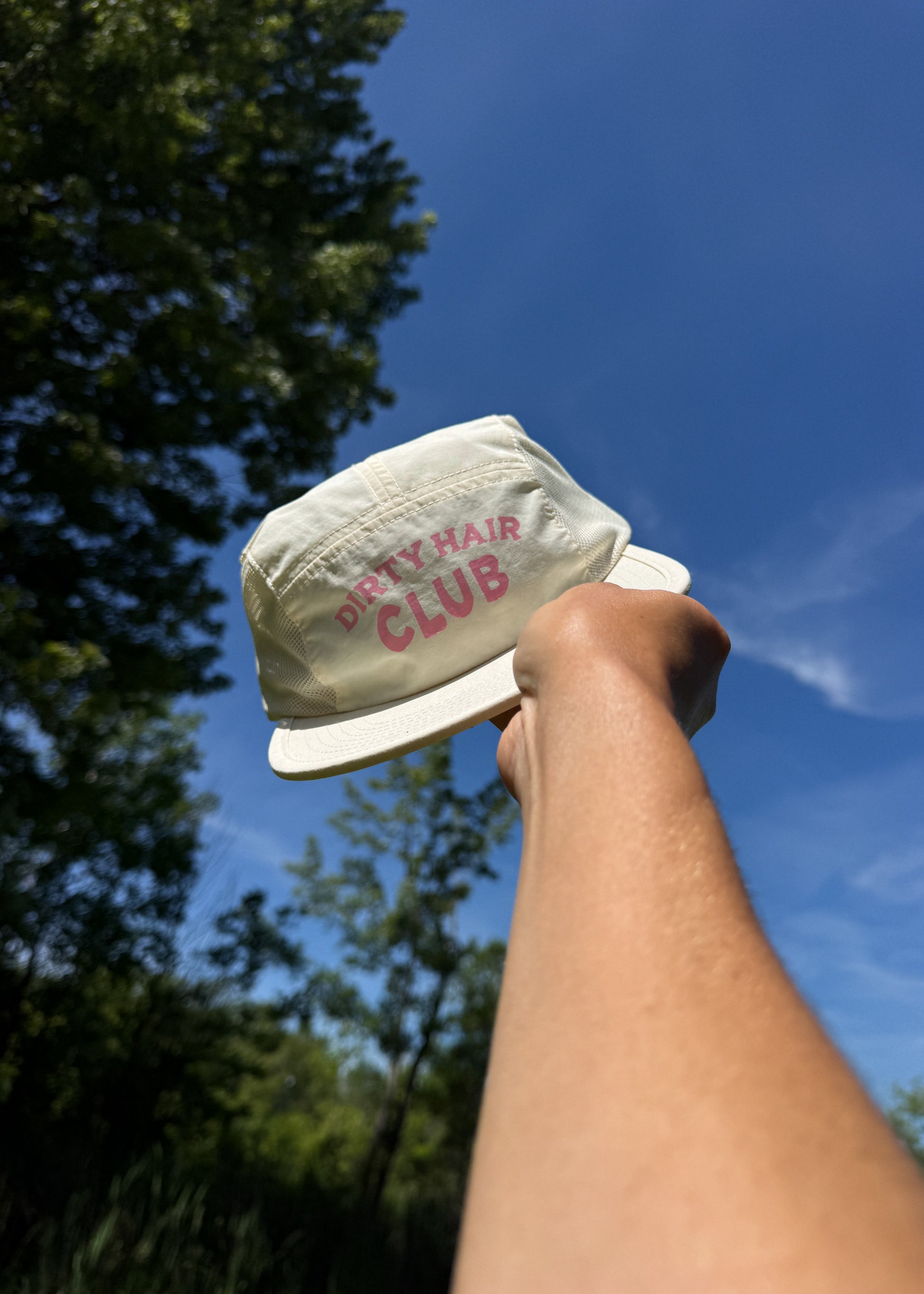 Hand holding a beige cap with 'Dirty Hair Club' pink text against a blue sky with trees