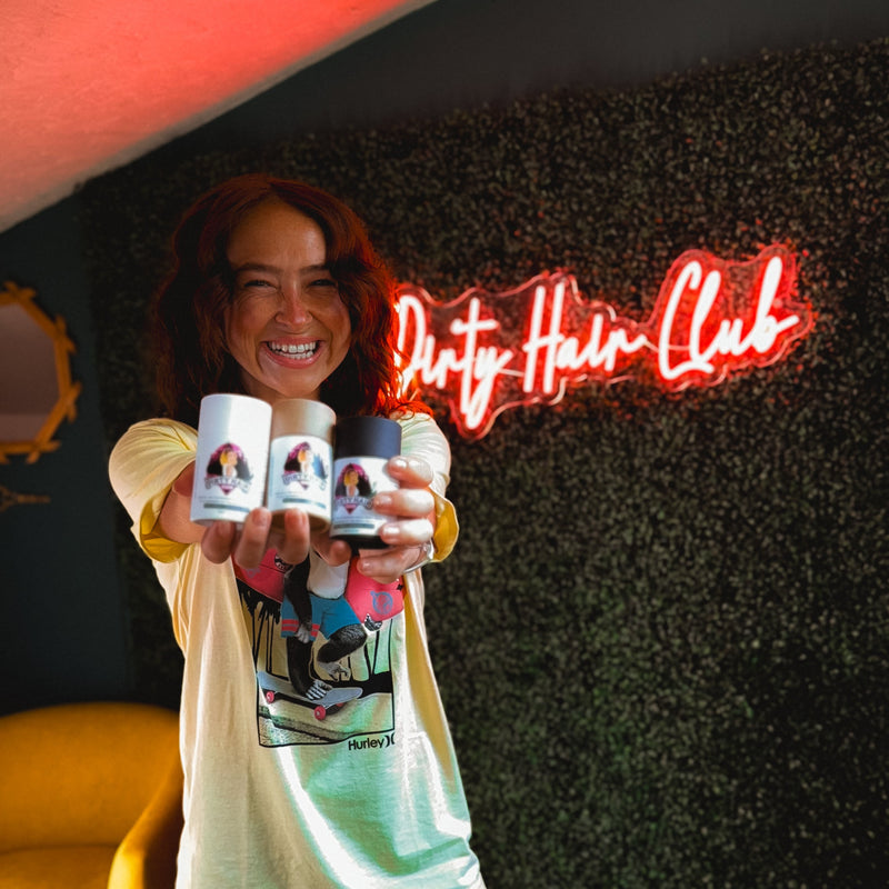 Person holding products with 'Dirty Hair Club' branding in a room with a neon sign.